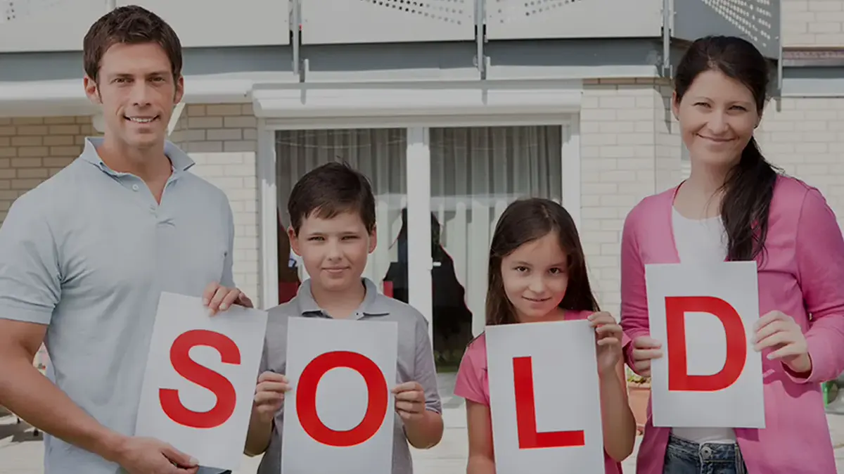 A smiling family holding up large letters that spell SOLD in front of their house
