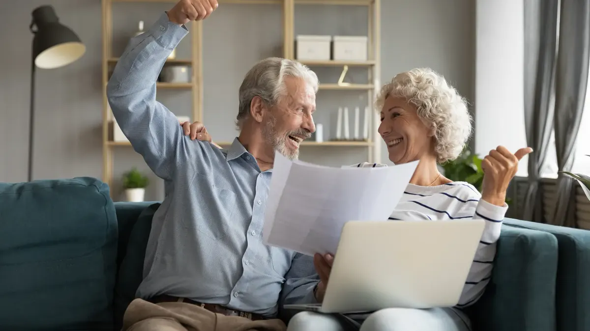 An older couple on a sofa smiling and cheering as they look at papers and a laptop
