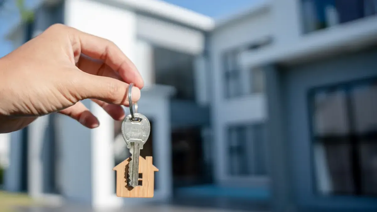 A hand holding up house keys with a house-shaped keyring in front of a new home
